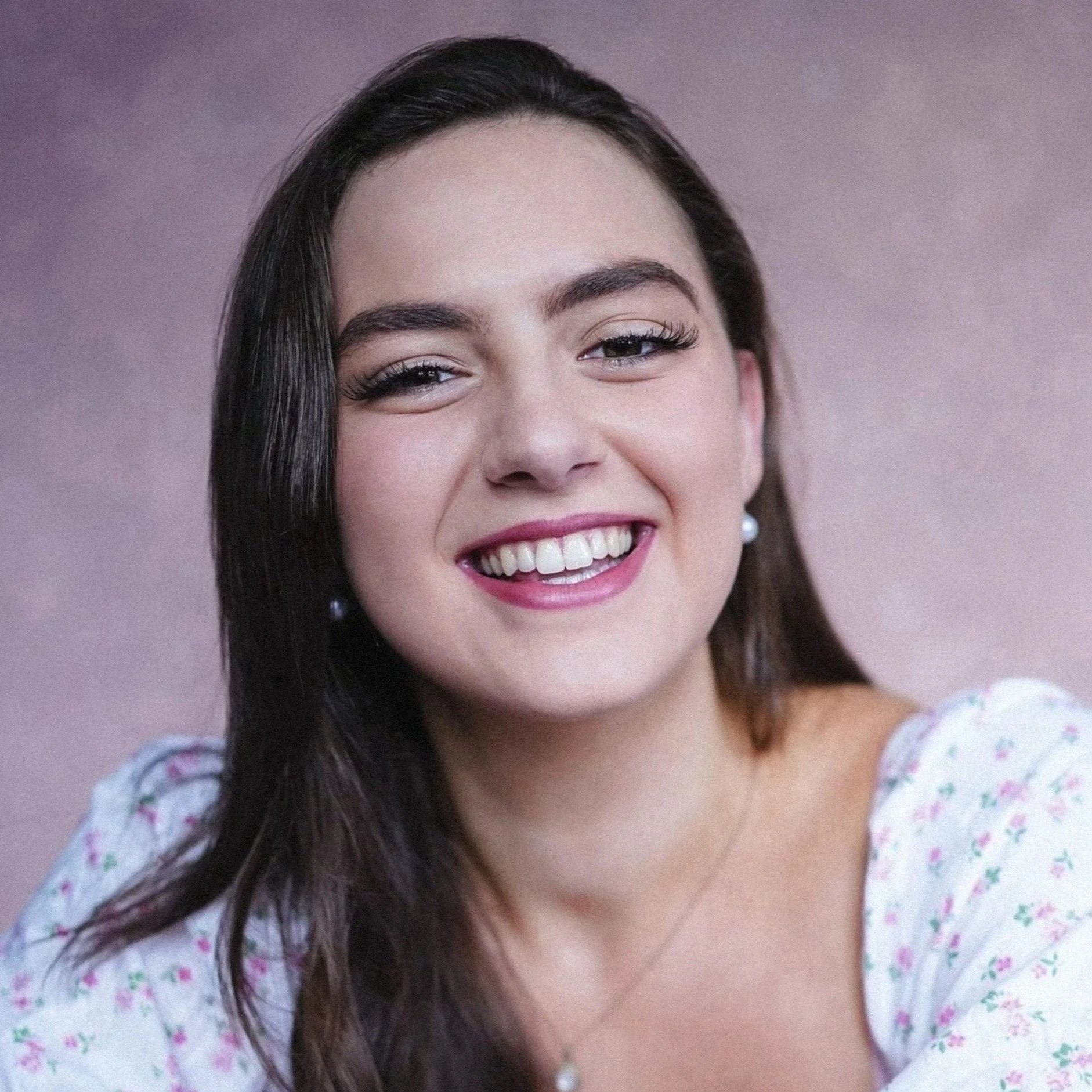 Close-up photo of a young woman with long dark hair smiling showing her teeth, wearing pearl earrings and a white top with pink floral pattern, against a pinkish background.