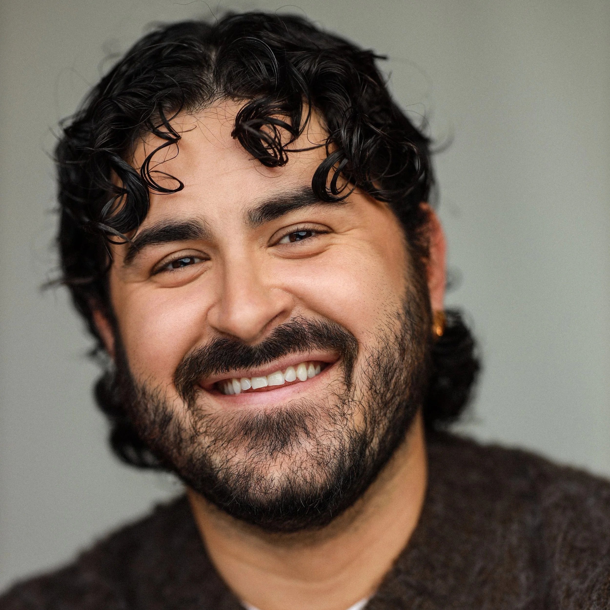 Close-up of a smiling man with curly dark hair and a beard, wearing a dark sweater, against a plain background.