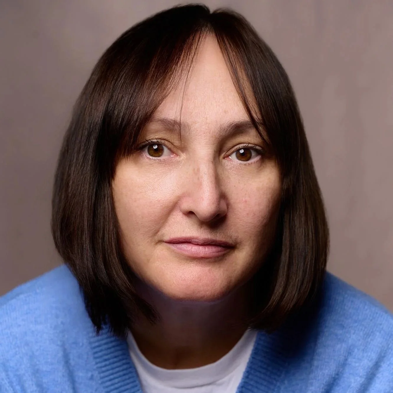 Close-up portrait of a woman with brown hair, wearing a blue sweater, against a neutral background.