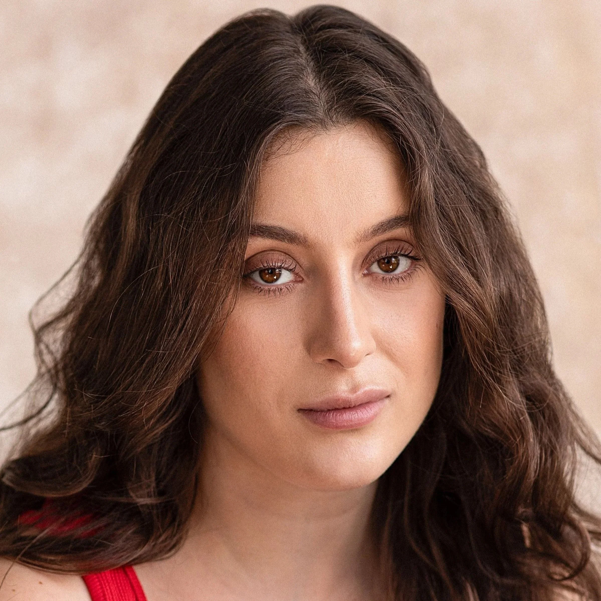 Close-up portrait of a woman with long, wavy brown hair and brown eyes, wearing a red top, against a neutral background.