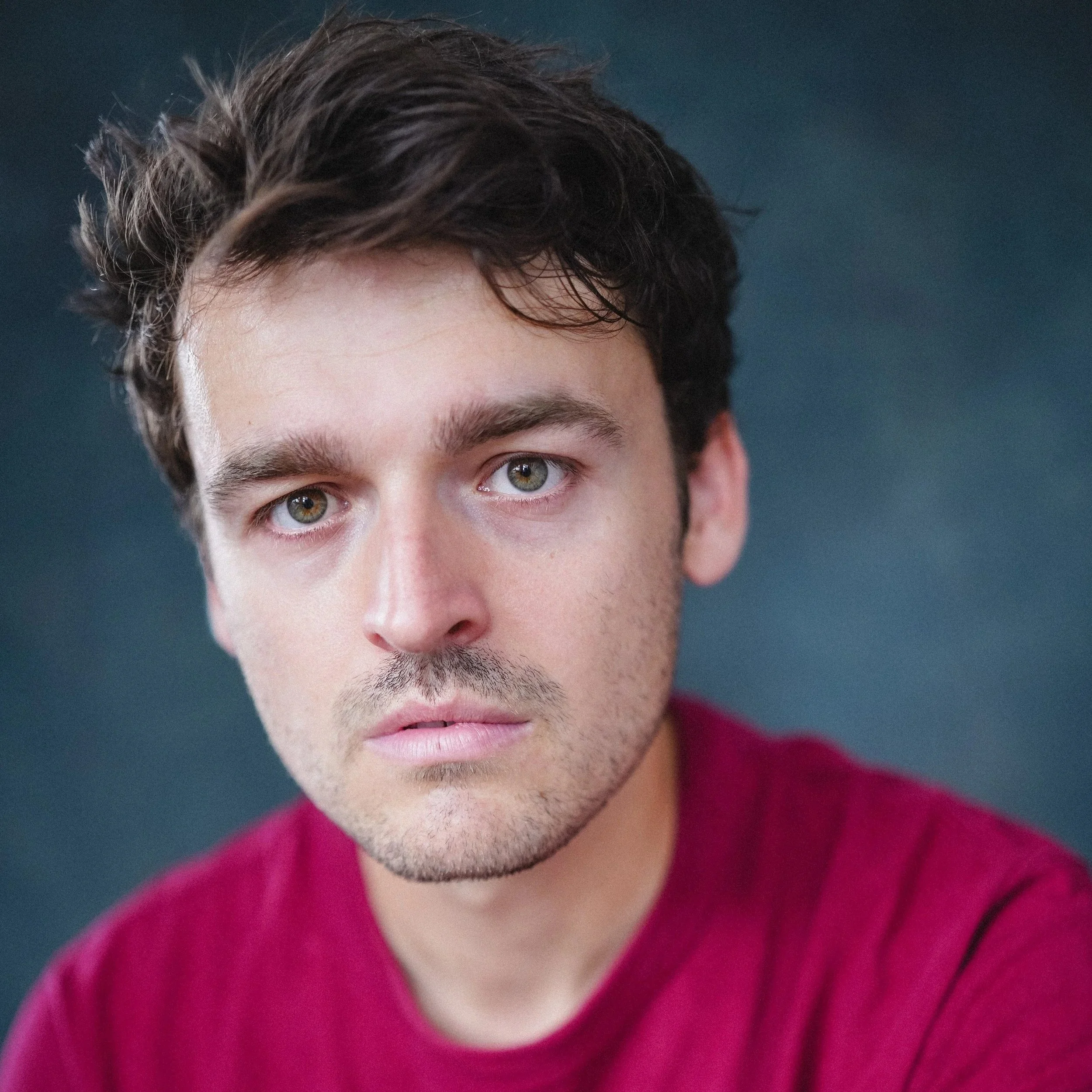 Close-up of a smiling young man with short dark hair, light skin, and hazel eyes, indoors with a blurred window in the background.