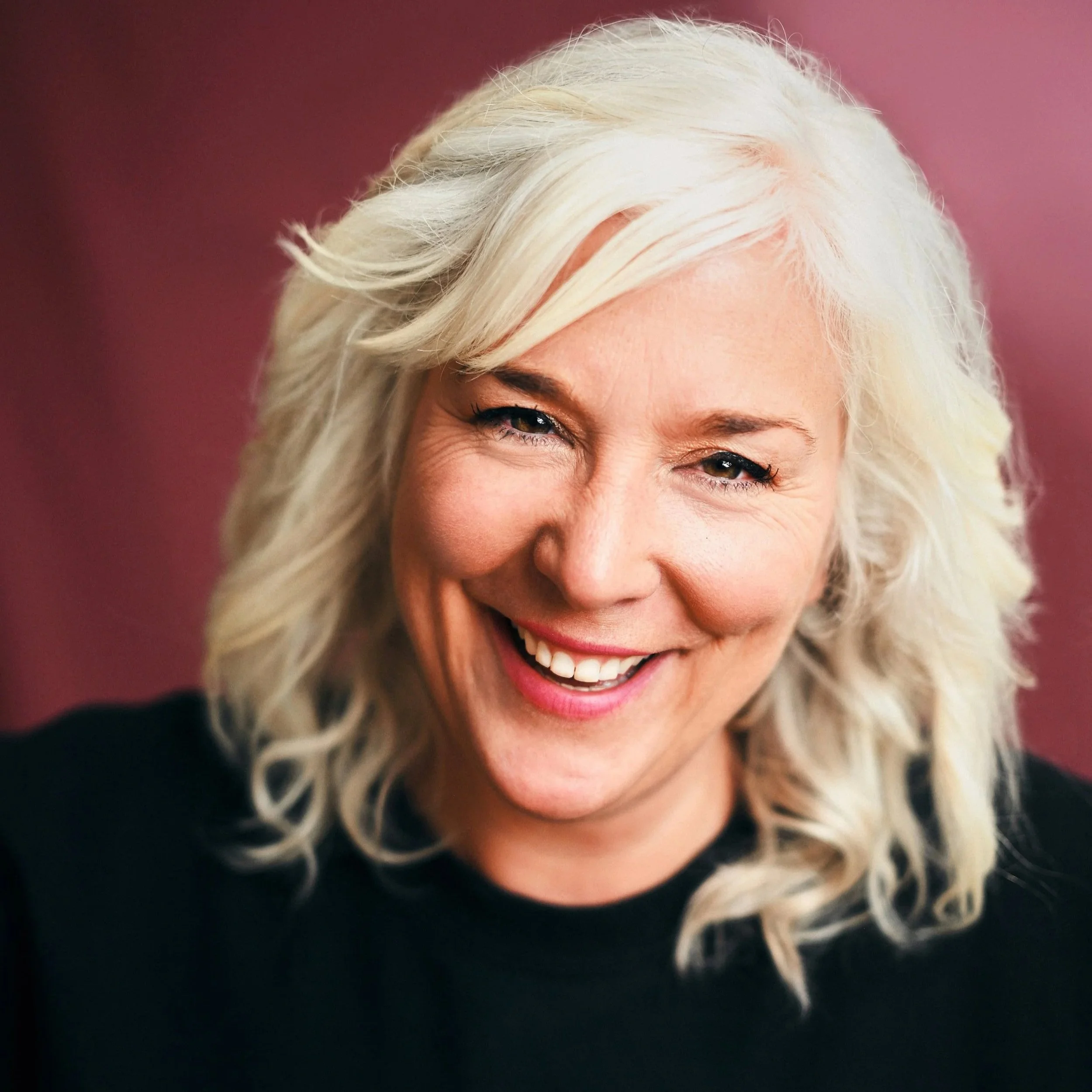 A smiling woman with wavy platinum blonde hair and light skin, wearing a black top, against a pink background.