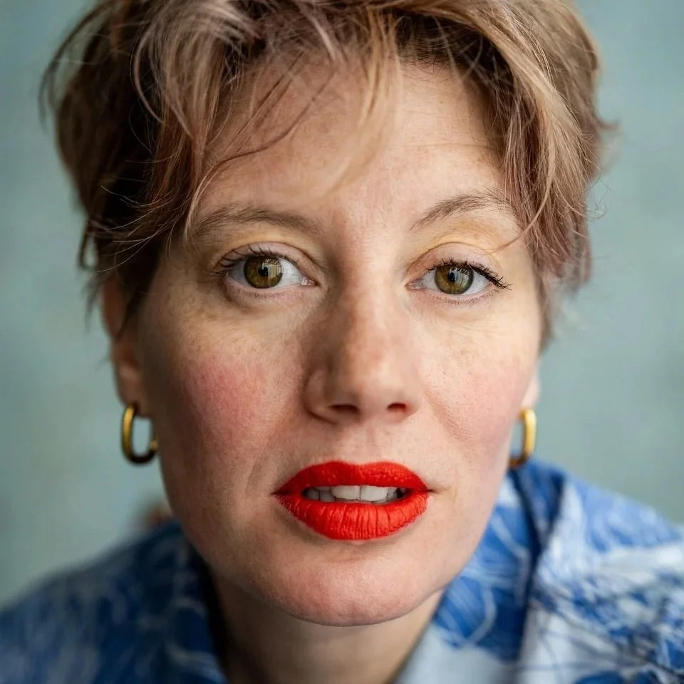 Close-up of a woman with short red hair, light skin, brown eyes, and wearing gold hoop earrings, with a neutral expression on her face against a purple background.