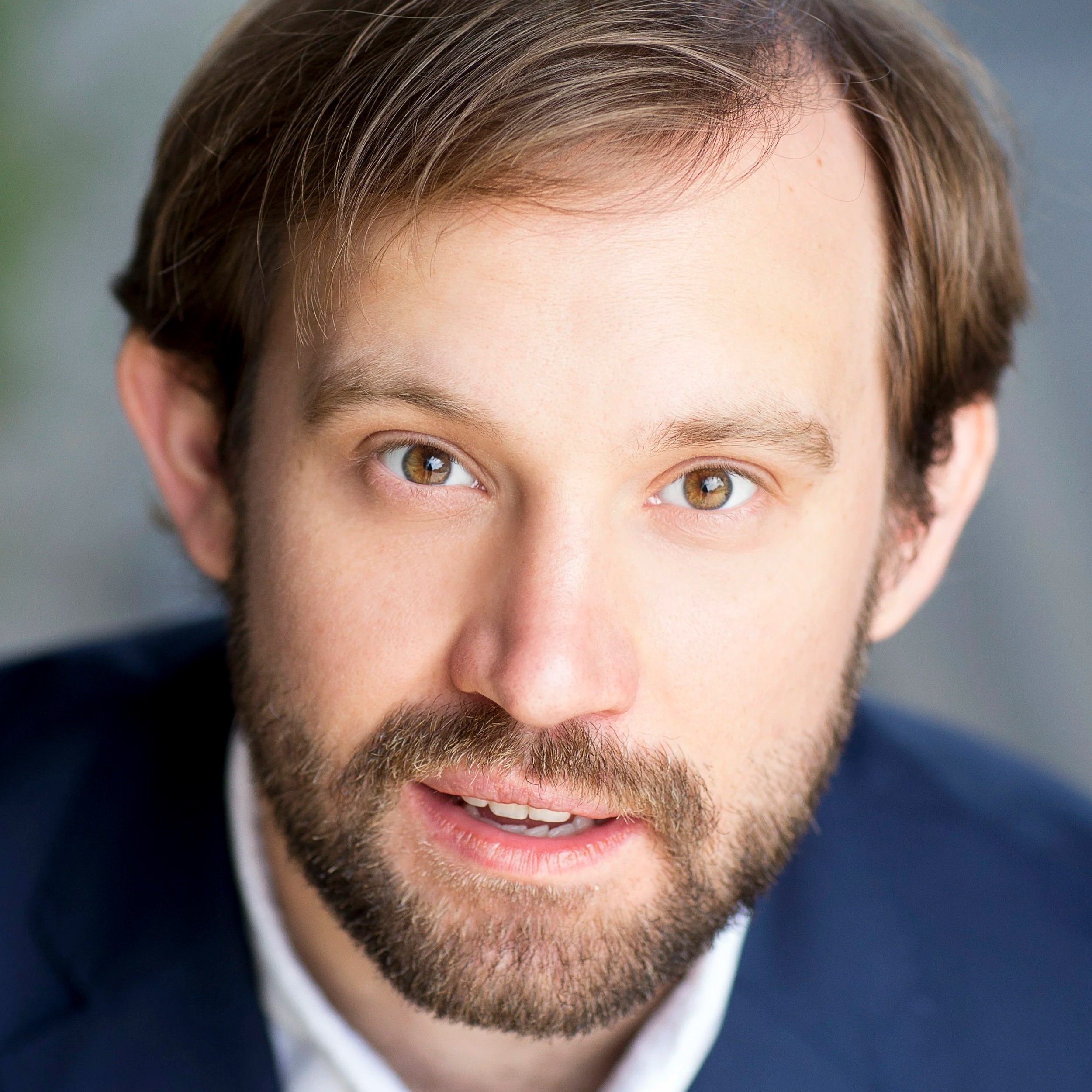 Close-up portrait of a young man with brown hair, brown eyes, and a beard, wearing a dark suit jacket and a white shirt, looking into the camera with a slightly open mouth.