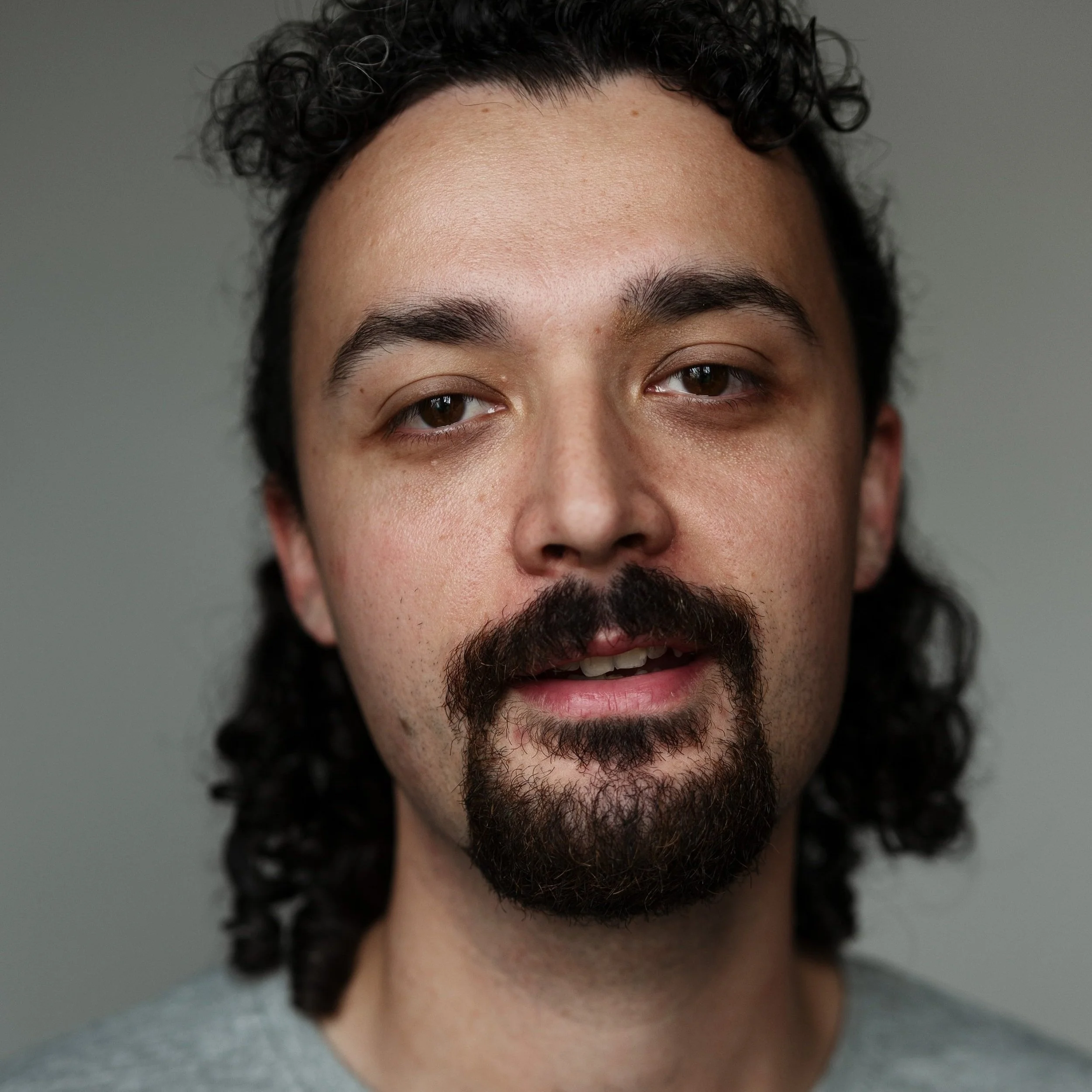 Close-up of a man's face with medium skin tone, dark curly hair, and a mustache and beard, looking at the camera.