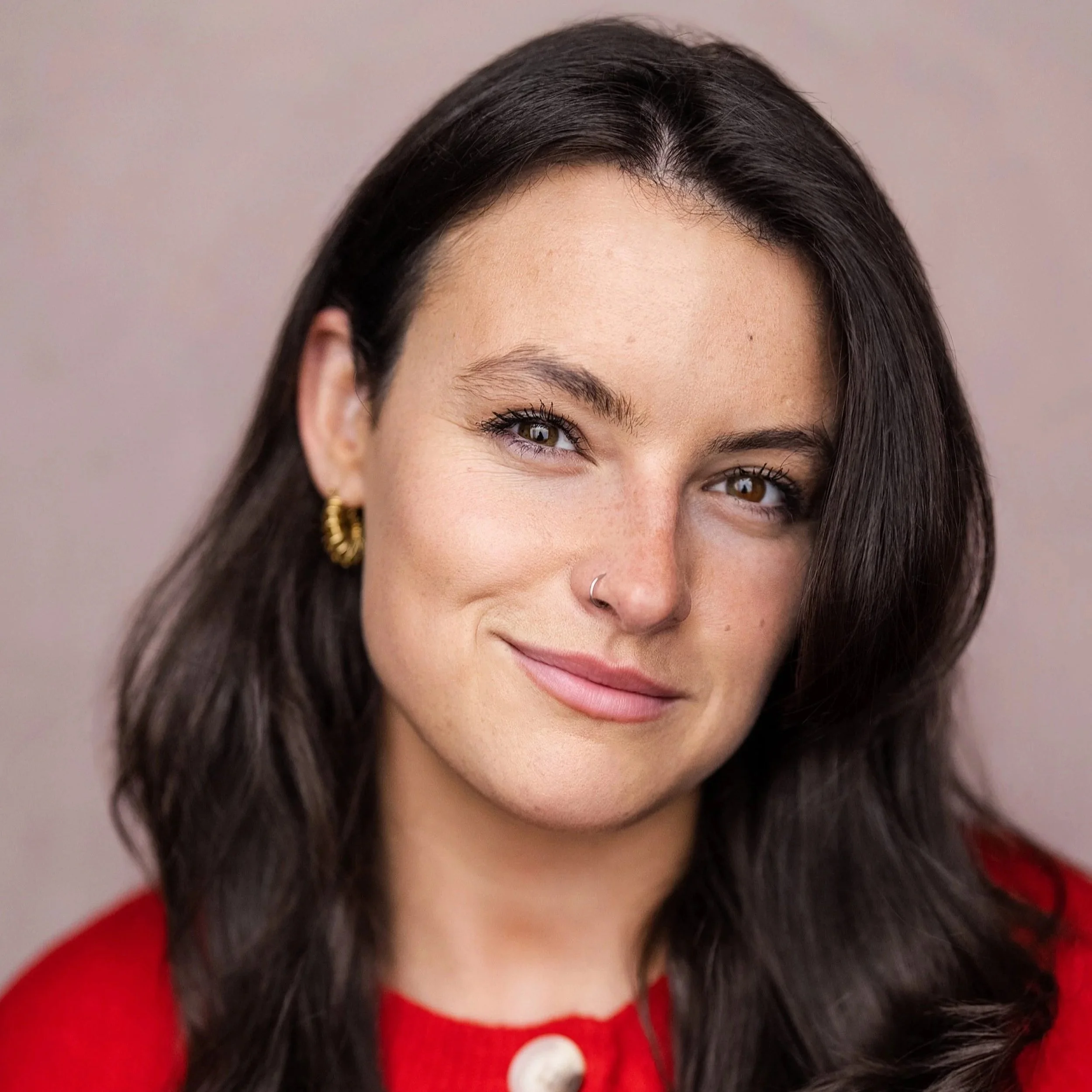 Close-up portrait of a woman with long dark hair, brown eyes, and a small hoop nose ring, wearing a red top against a gray background.