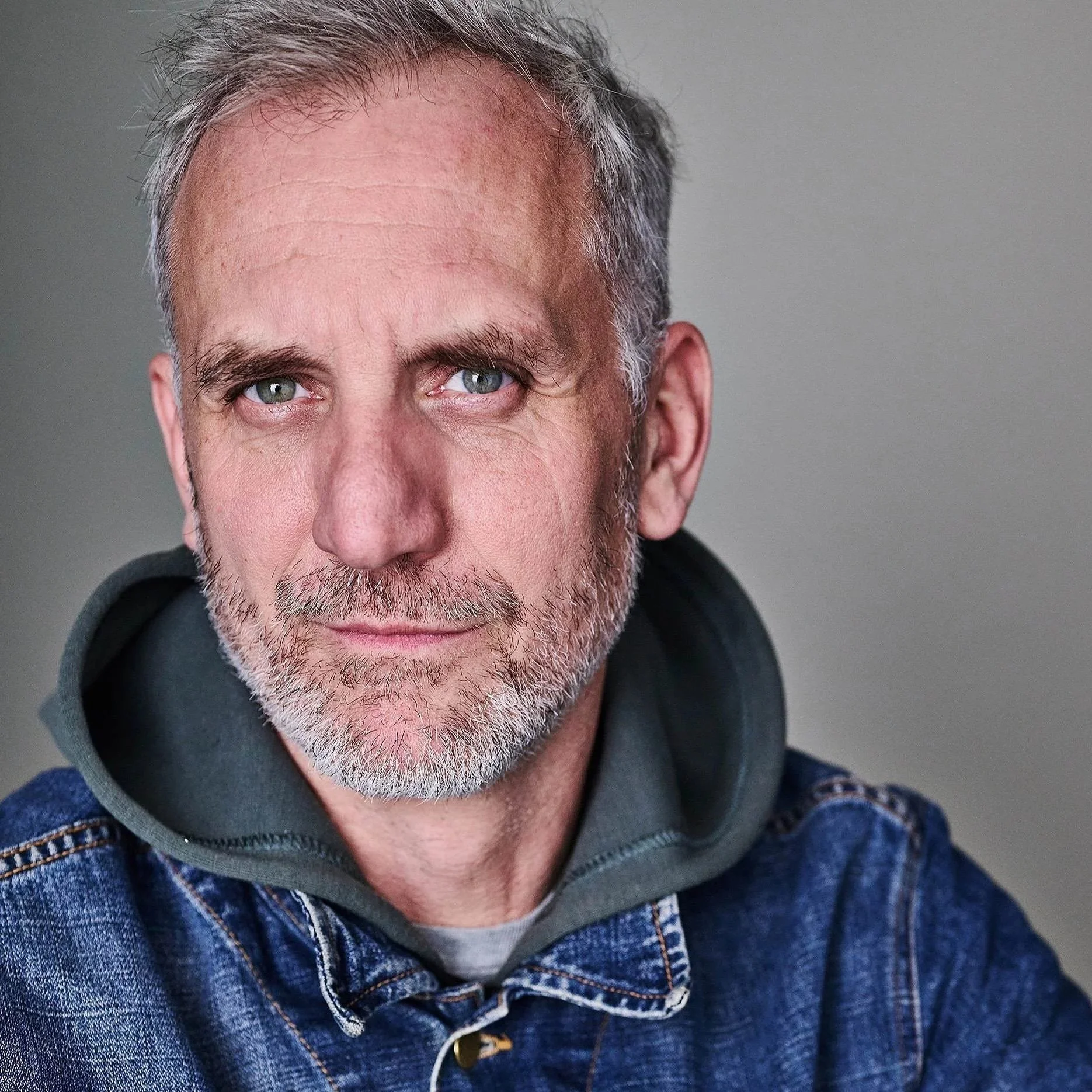 Close-up of a middle-aged man with gray hair, blue eyes, and a beard, wearing a denim jacket over a gray hooded sweatshirt, facing the camera.