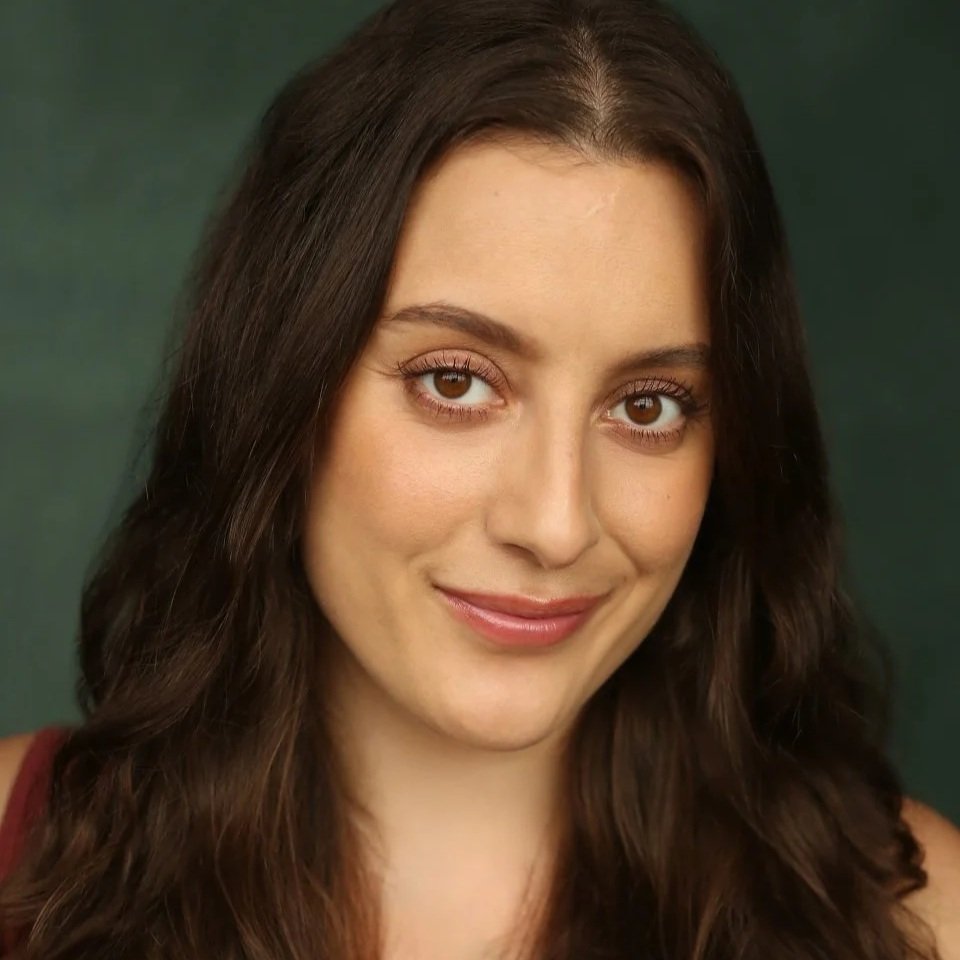 Close-up portrait of a woman with long, wavy brown hair and brown eyes, wearing a red top, against a neutral background.