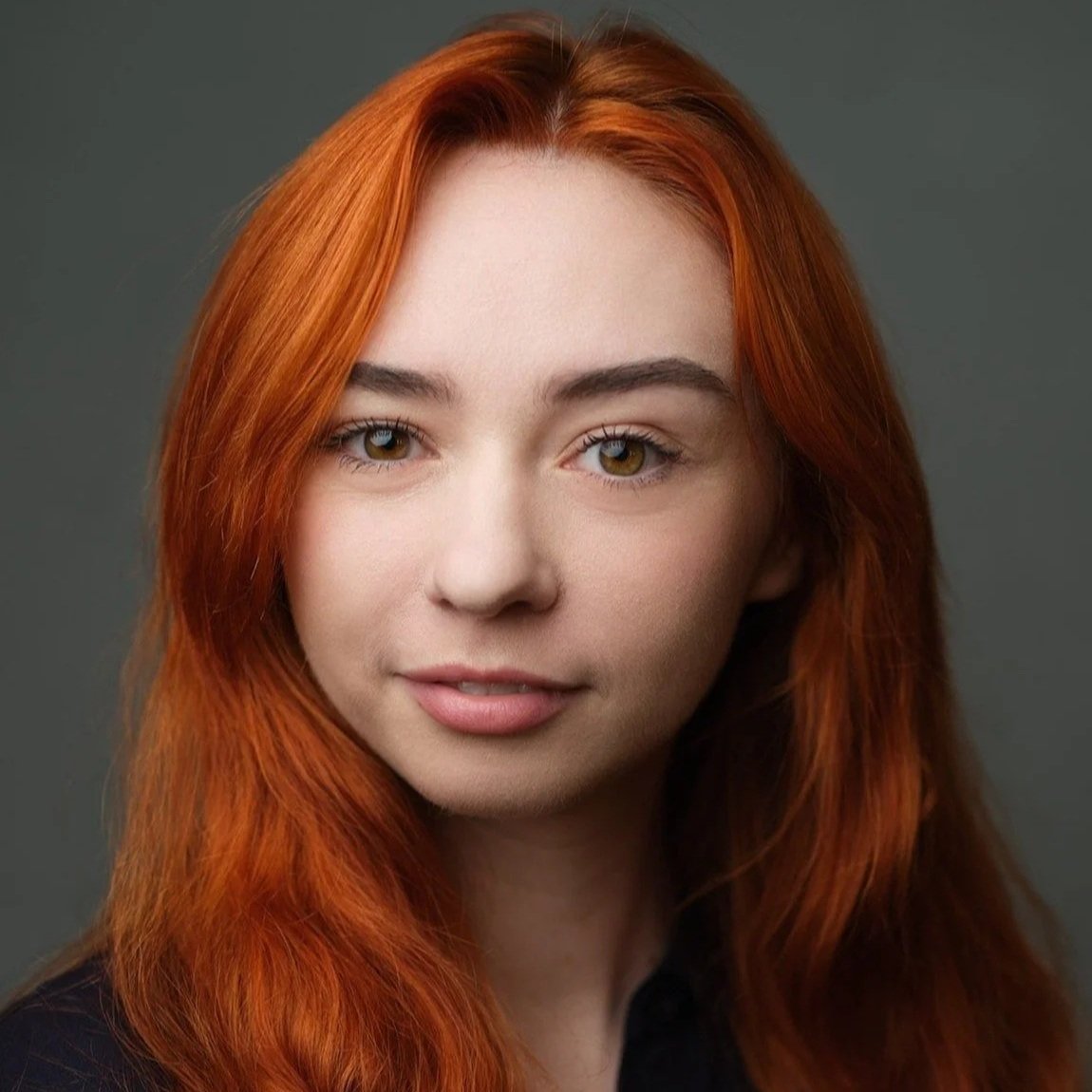 A young woman with long, straight red hair wearing a black blazer against a dark background.