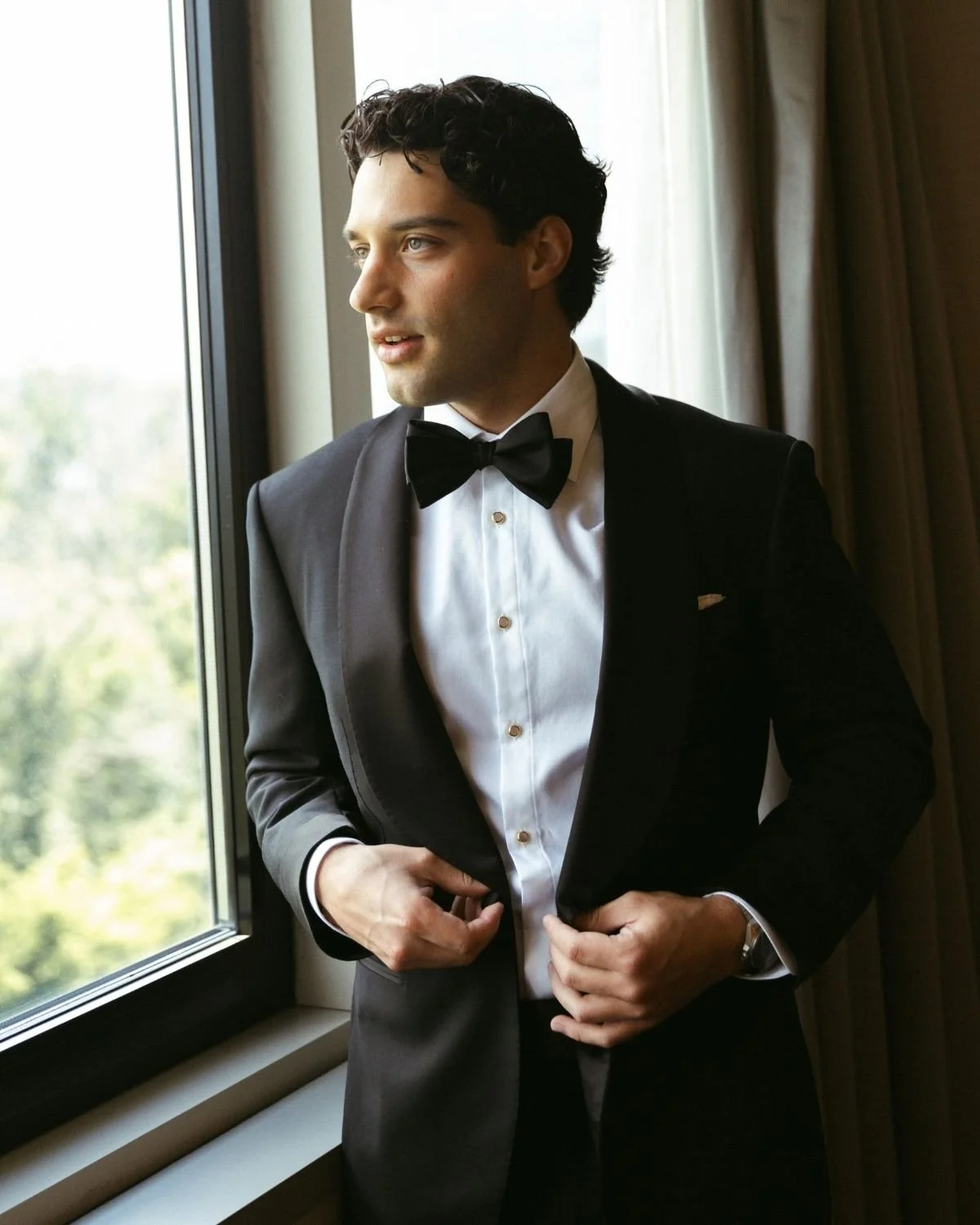 A groom in a black shawl collar tuxedo by a window