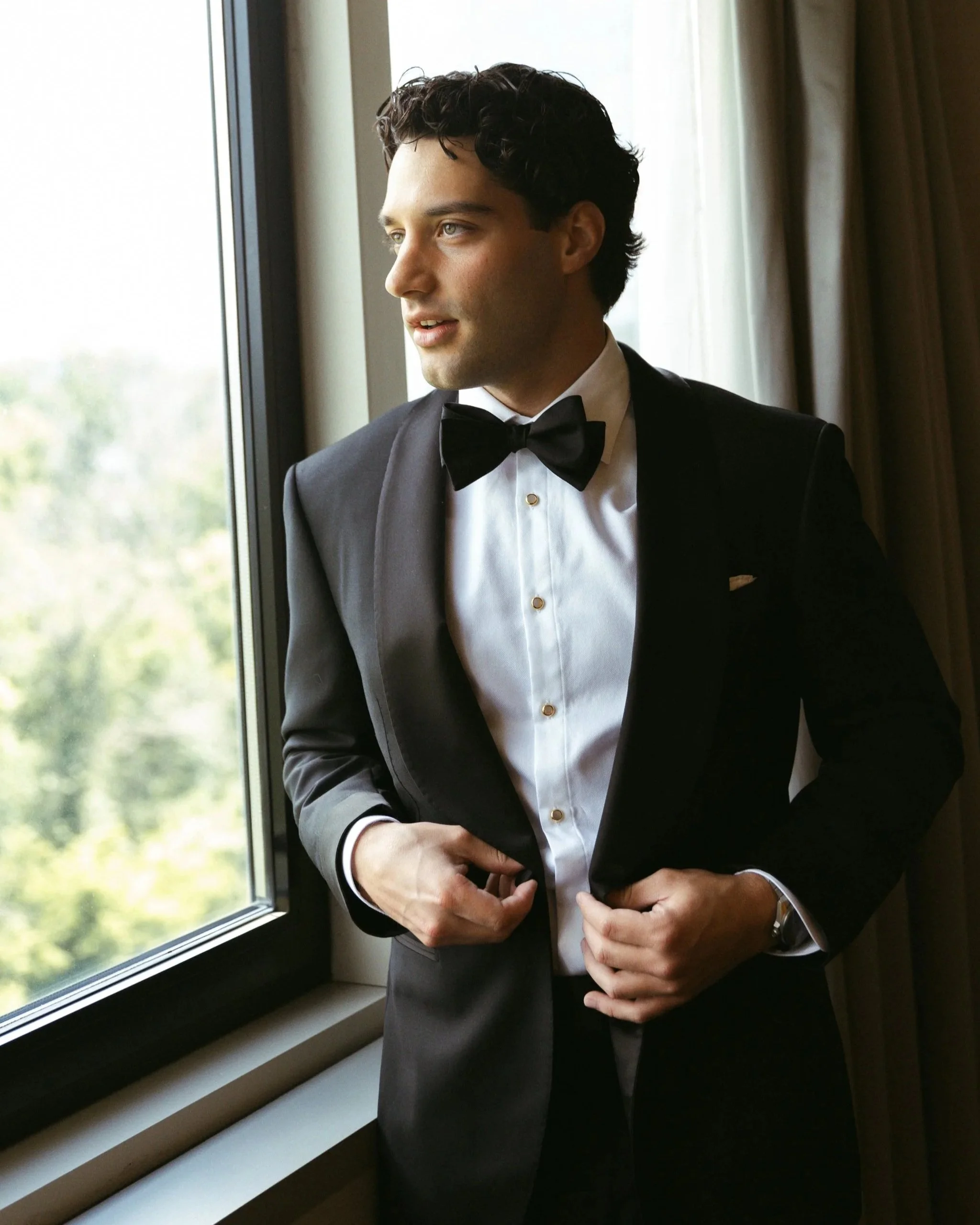 A groom in a black shawl collar tuxedo by a window