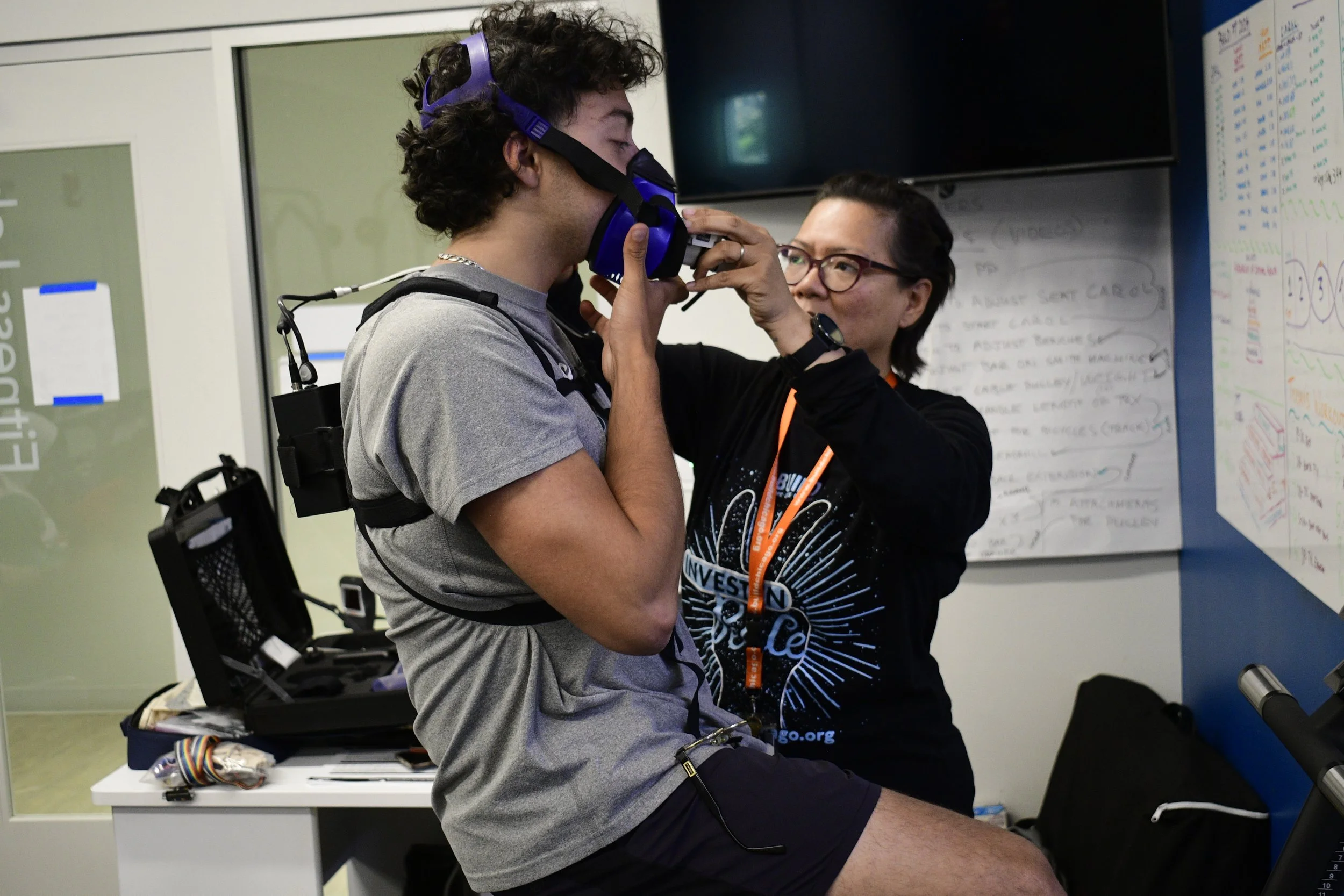 A person assists another person wearing a VR headset and harness in a room with equipment and notes on a whiteboard.