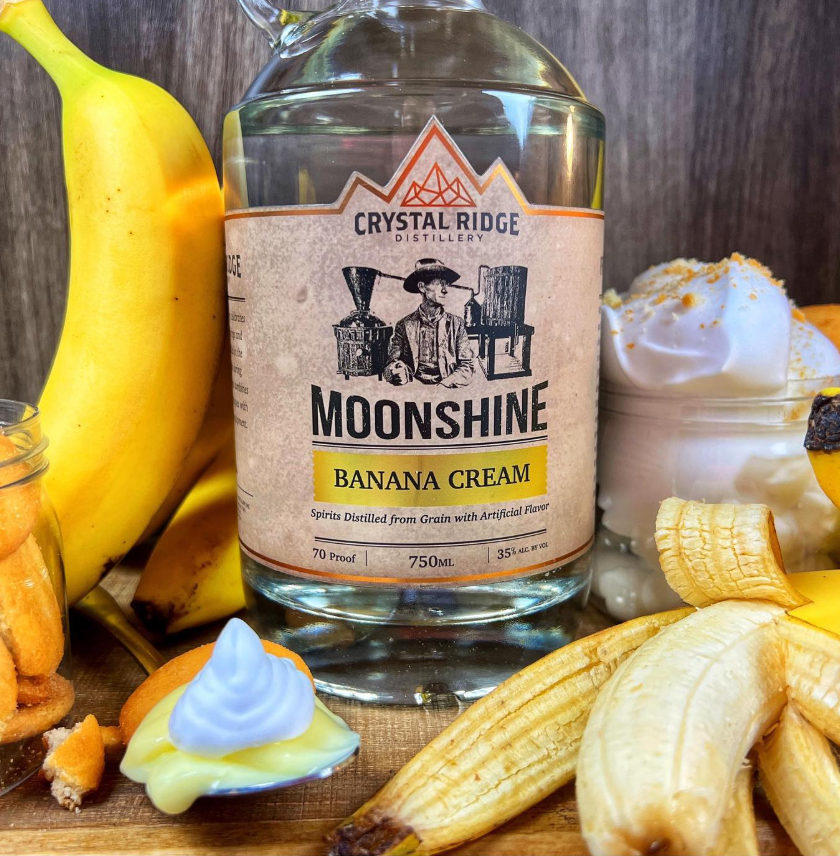 A bottle of Crystal Ridge Moonshine Banana Cream with ripe bananas around it, a spoon with banana pudding, and a bowl of whipped cream with banana slices.