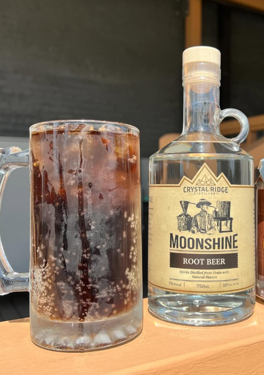A glass mug filled with a dark, fizzy root beer with ice, next to a clear bottle labeled 'Moonshine Root Beer' from Crystal Ridge Distillery.