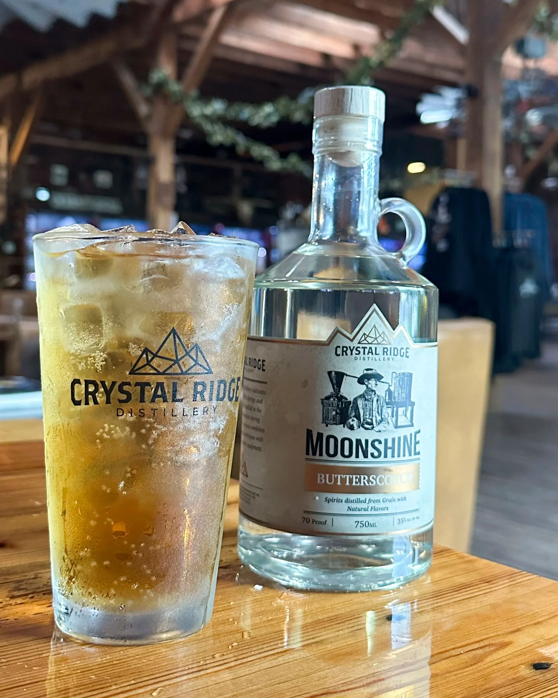 A glass of cold beverage with ice and condensation, with the label 'Crystal Ridge Distillery' and an illustration, next to a clear bottle of Moonshine Butterscotch liquor on a wooden table inside a rustic setting.