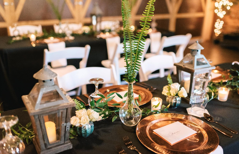 Elegant table setting with black tablecloth, gold chargers, white chair, glass vases with greenery, candles, lanterns, and place cards.