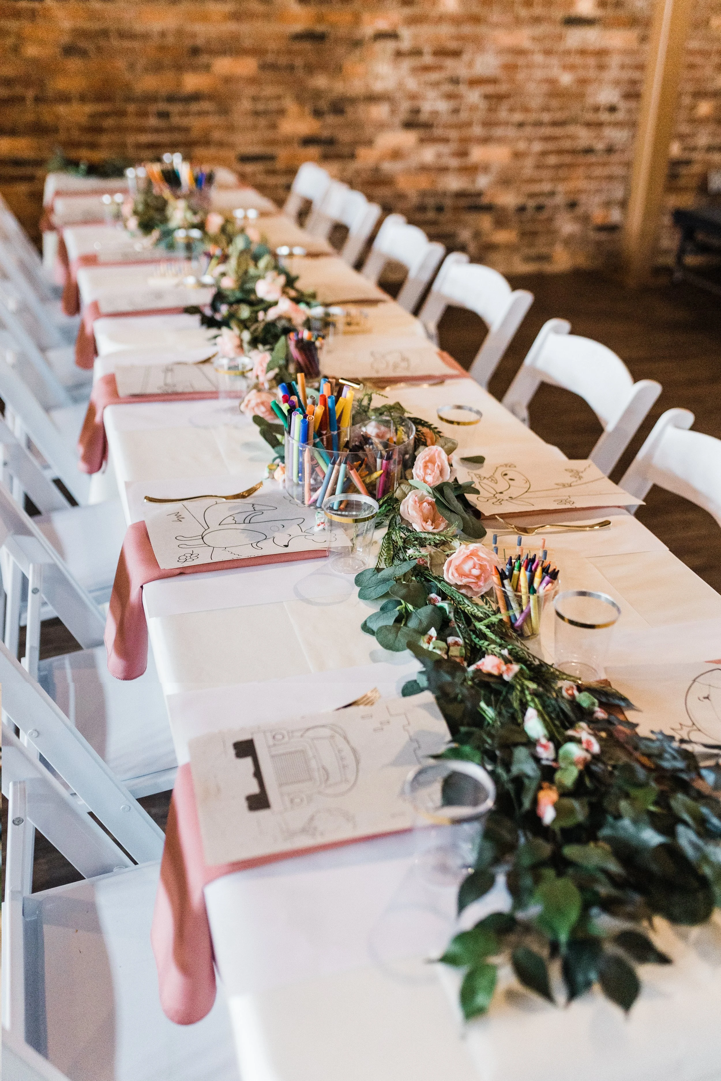 Long rectangular table decorated with a floral garland, coloring pages, and cups filled with colored markers, set for a celebration or event.