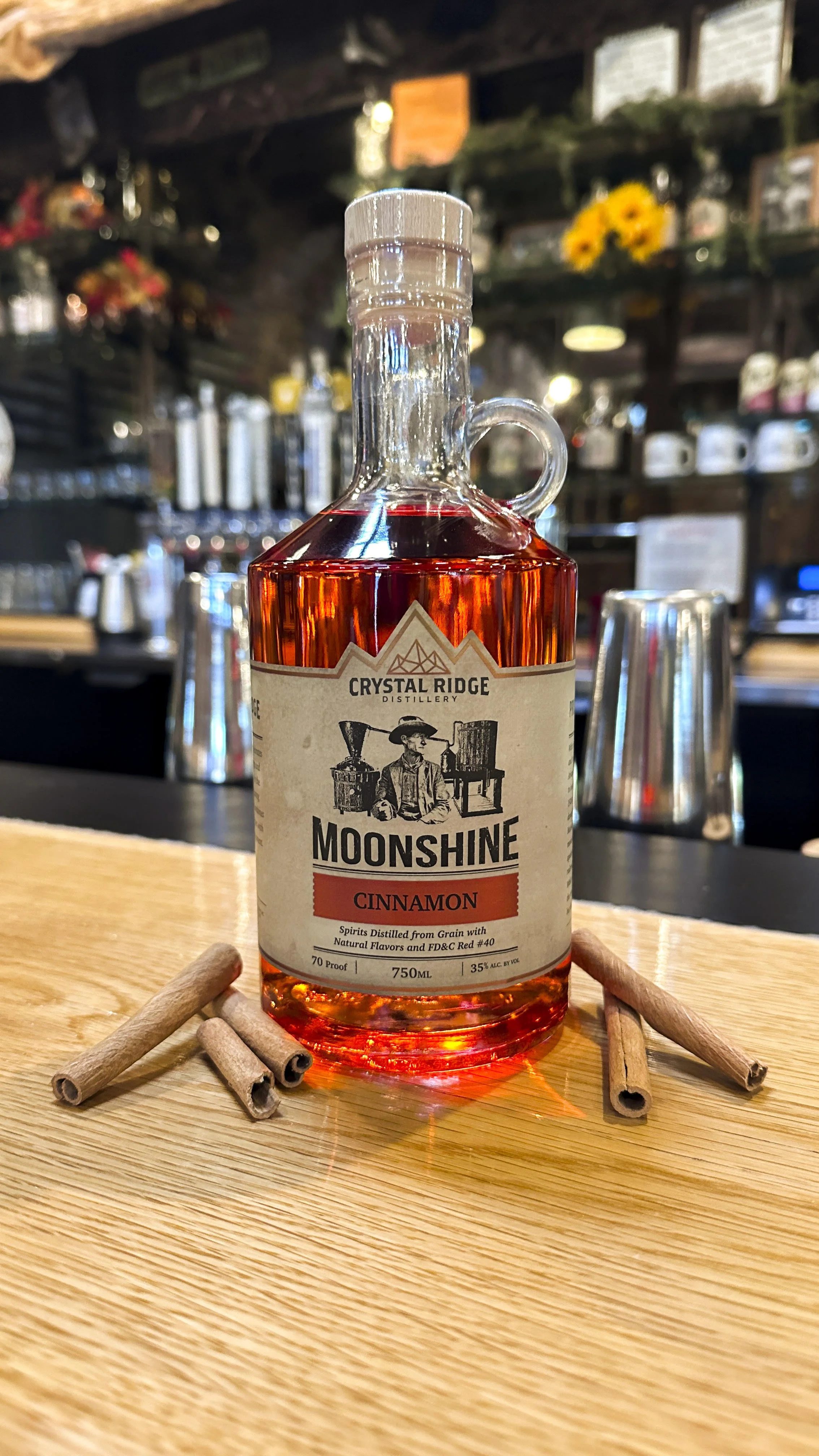 A bottle of Moonshine Cinnamon on a wooden surface surrounded by cinnamon sticks, with a blurred bar background.