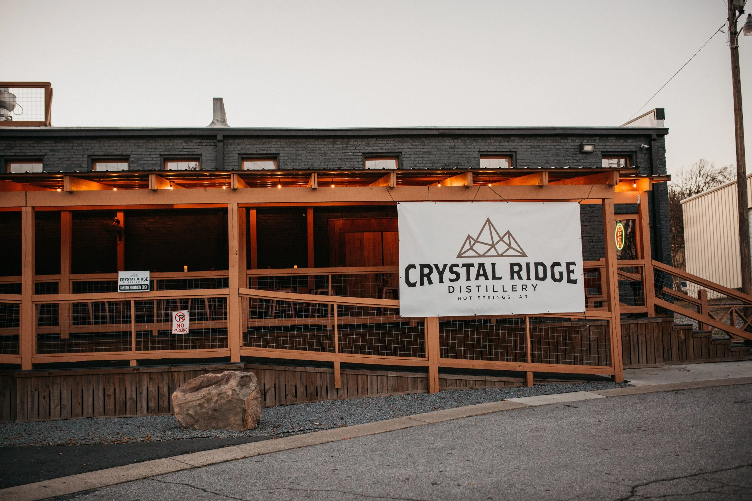 Exterior of Crystal Ridge Distillery in Hot Springs, Arkansas, showing a large sign with a mountain logo and the name, with a wooden balcony, gravel pathway, and a large rock in front.