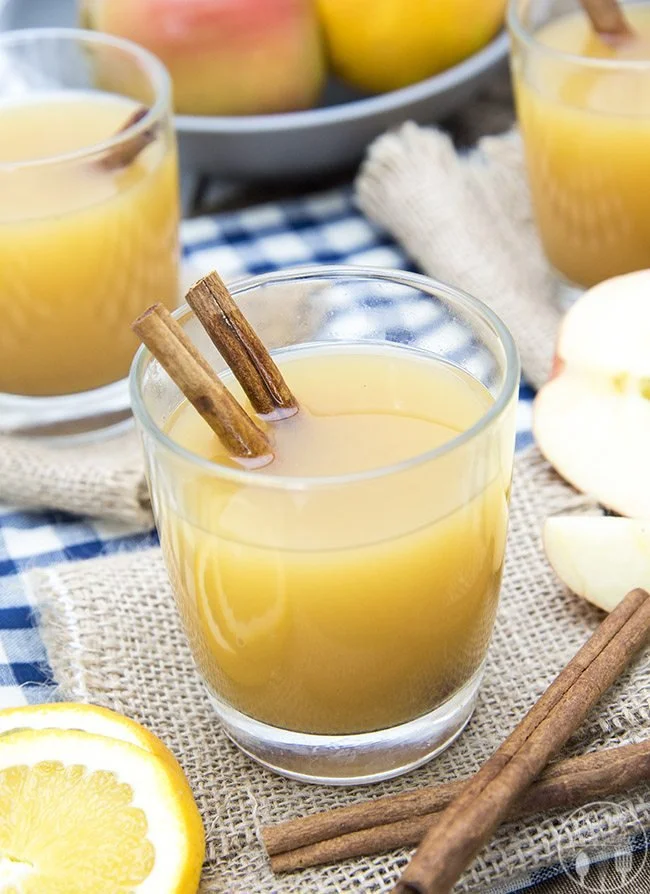 A glass of apple cider garnished with two cinnamon sticks, on a rustic table with fresh apples, orange slices, and more glasses of cider in the background.
