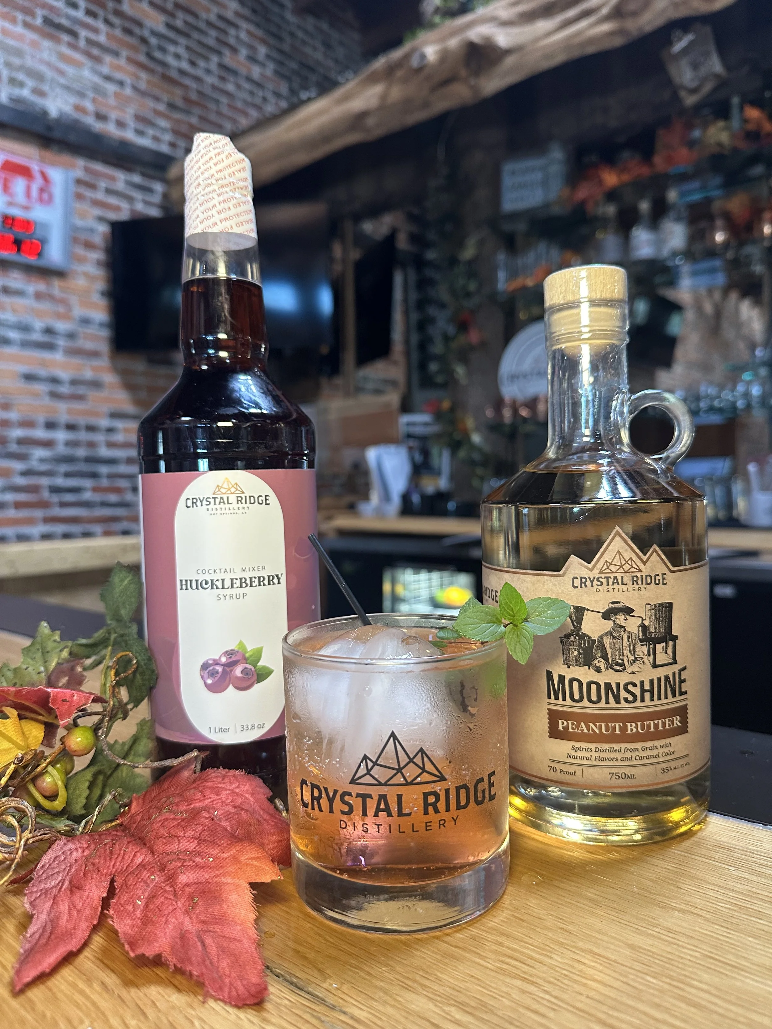 A cocktail in a Crystal Ridge Distillery glass garnished with a mint sprig, beside bottles of Moonshine Peanut Butter and Huckleberry Syrup, with autumn leaves and berries on a wooden surface.