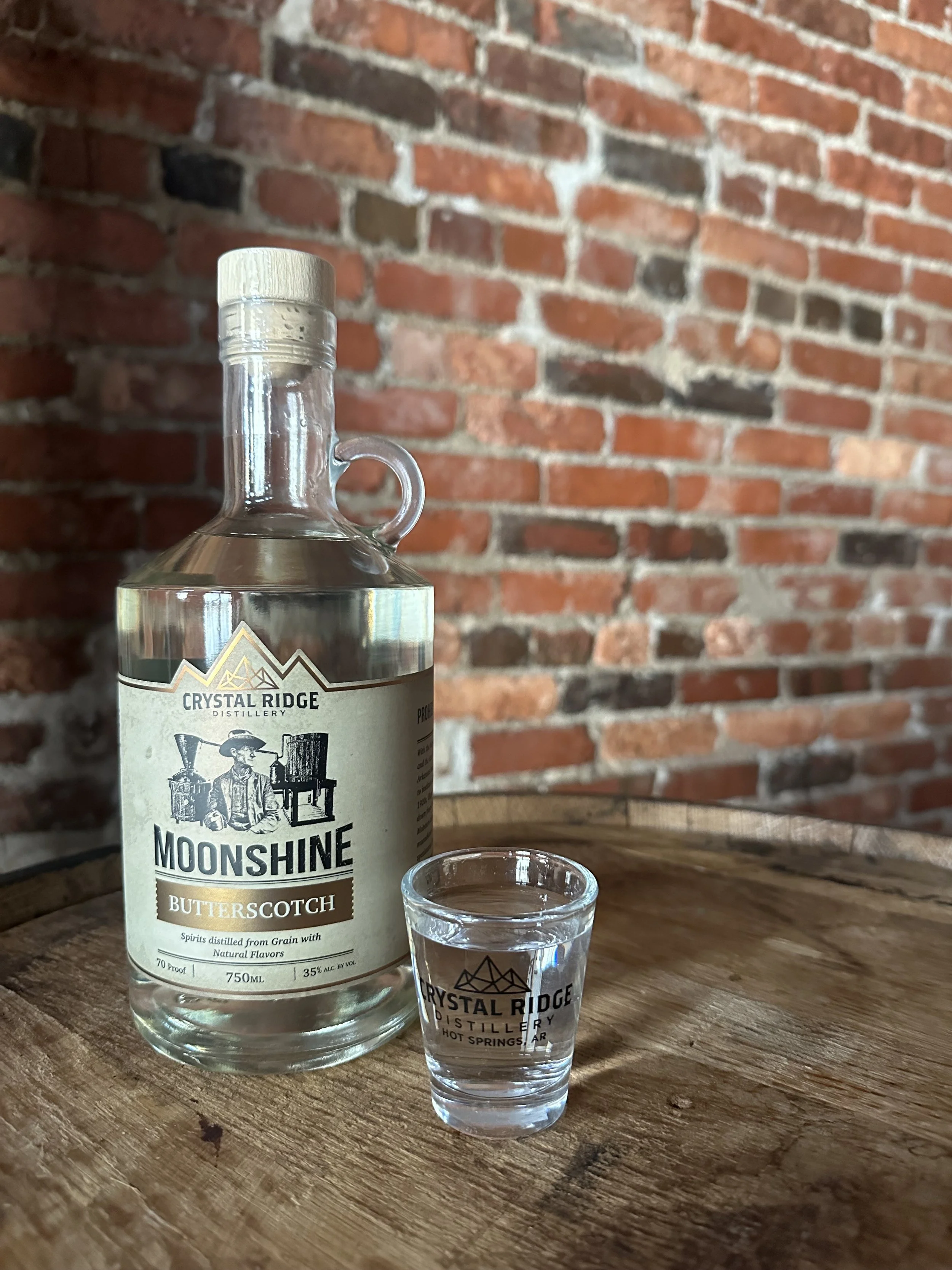 A clear glass bottle of Moonshine Butterscotch liquor and a small shot glass with the Crystal Ridge Distillery logo, both placed on a wooden surface with a brick wall background.