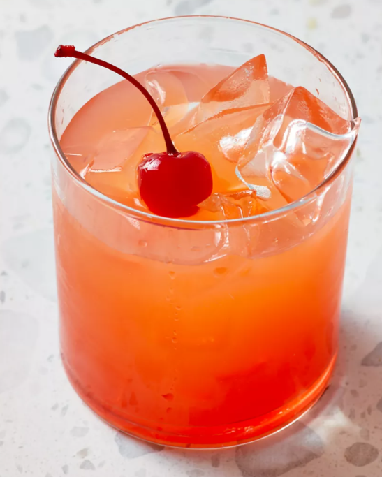 A glass of pink cocktail with ice cubes and a maraschino cherry garnish on top.
