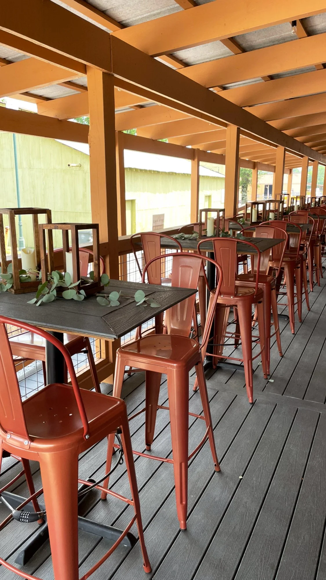 Outdoor patio dining area with rust-colored metal barstools, black wooden tables decorated with lanterns, candles, and greenery, surrounded by a wooden railing. The back patio also has bistro lighting for nice ambiance.