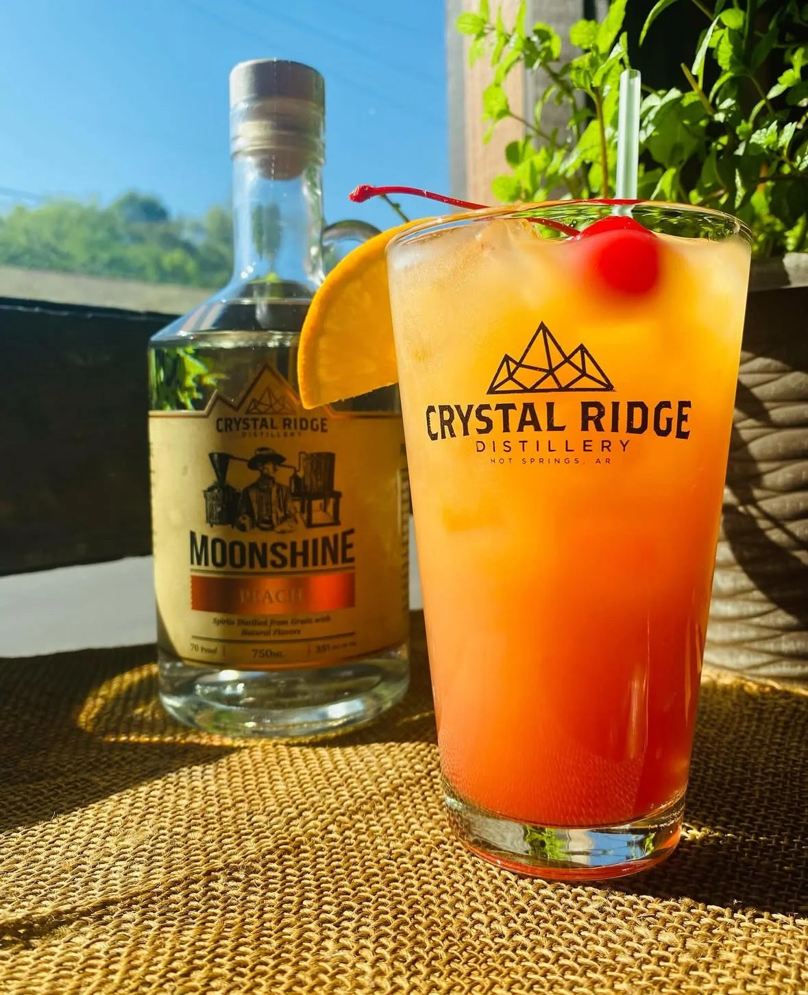 A colorful cocktail with a cherry and lemon wedge in a Crystal Ridge Distillery glass, with a bottle of Moonshine Peach behind it, set on a woven mat outdoors with sunlight and greenery in the background.