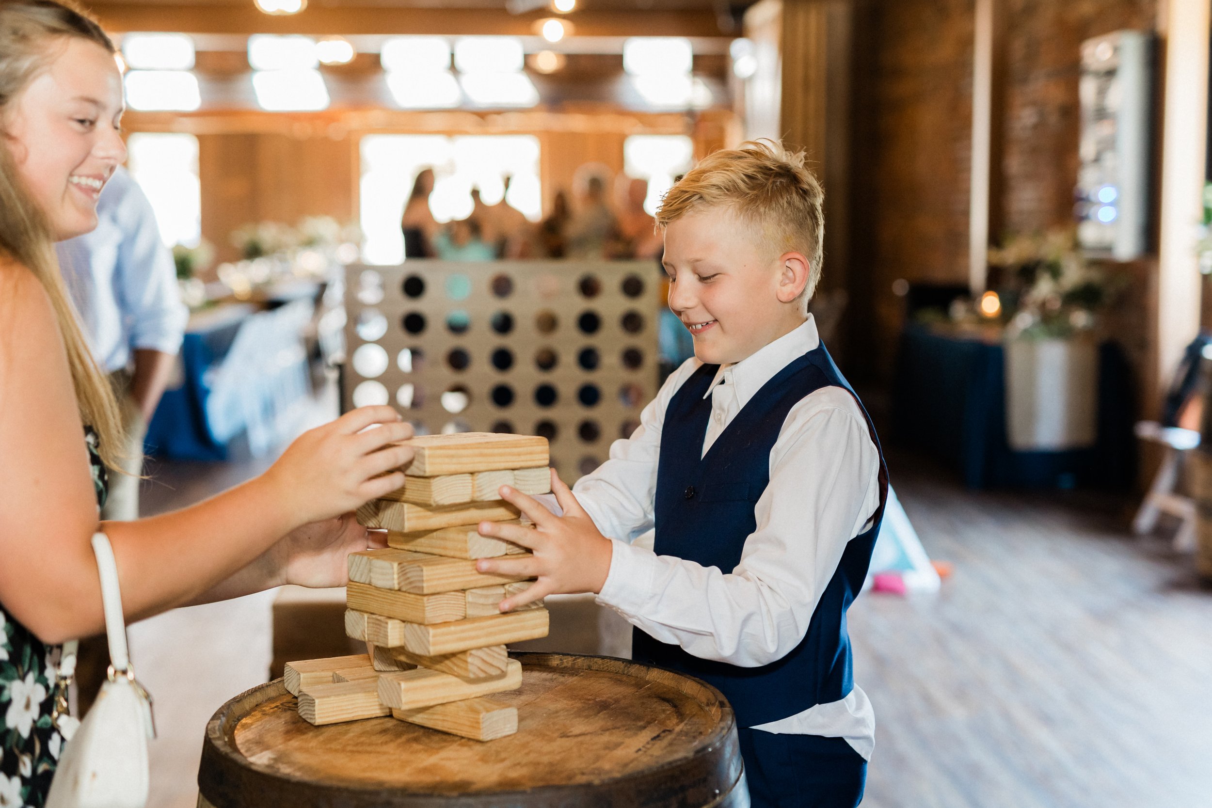A young boy and girl playing a game of giant Jenga at a social gathering, indoors with wooden walls and blurred background guests. Considering renting our family-friendly games to keep all ages occupied during the visit.