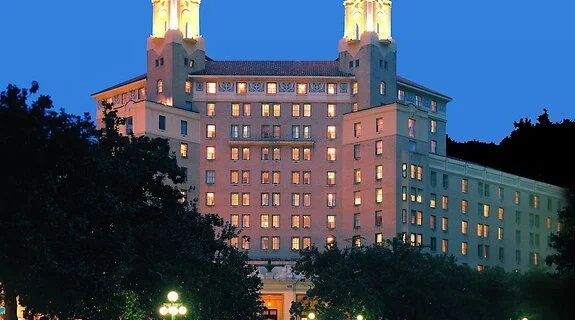 A large hotel or building with towers at the top, illuminated against a blue evening sky, with trees in the foreground.
