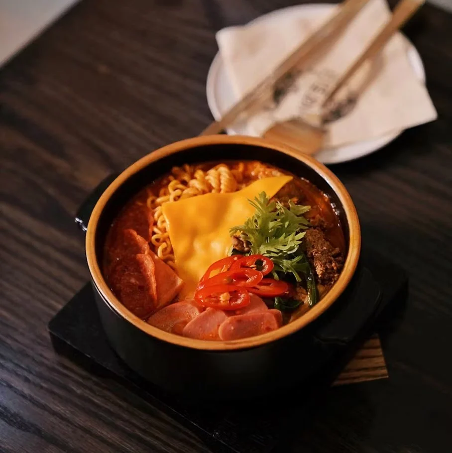 Tiger budae jjigae — Korean spicy stew with kimchi, spam, assorted sausages, ramen noodles, and cheese
