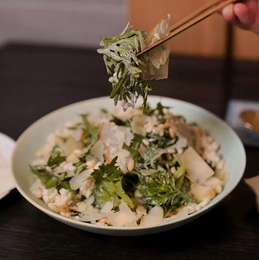 Chrysanthemum salad dressed in black sesame sauce with pear slices and crispy puffed rice, lifted by chopsticks above the bowl