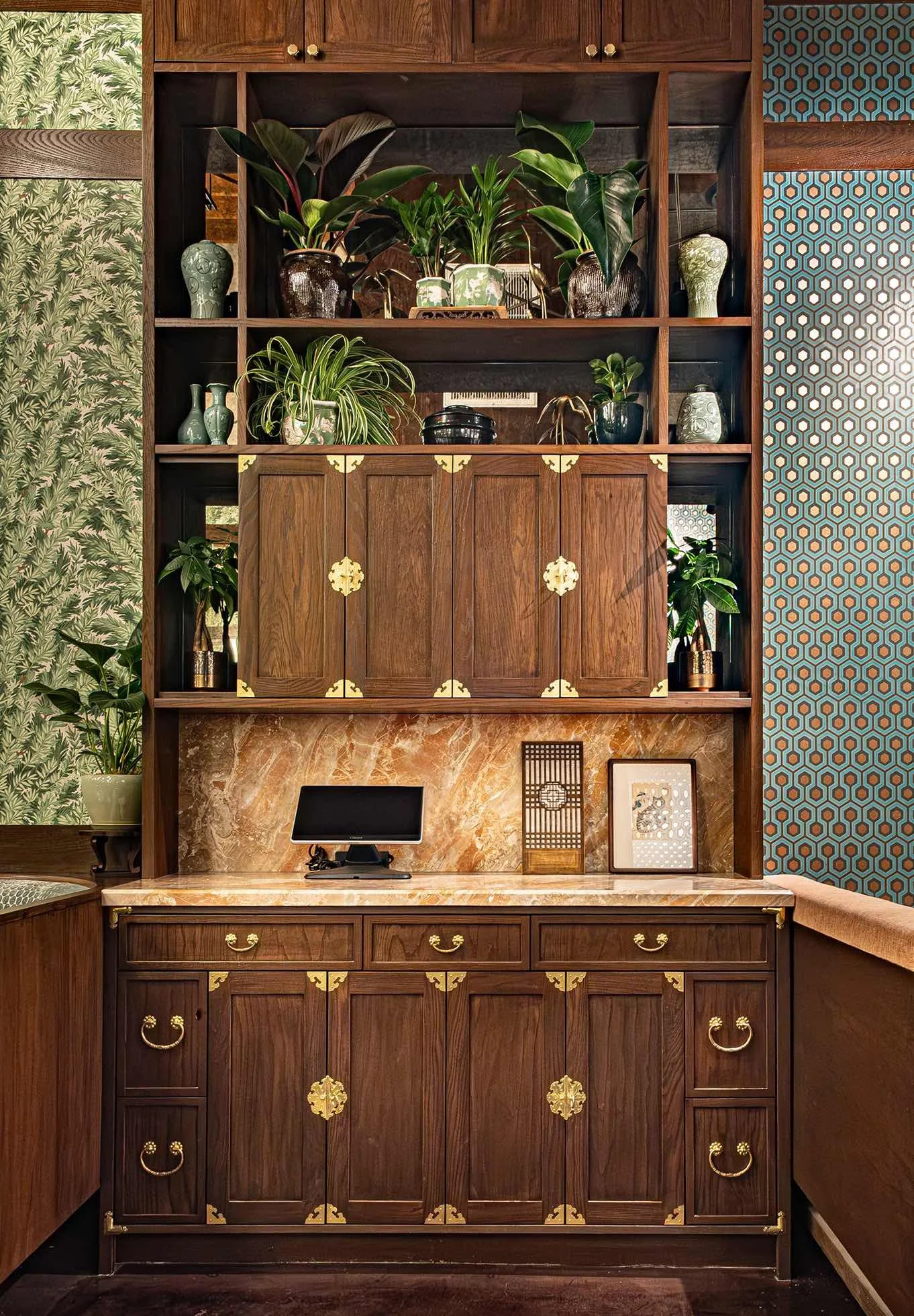 Restaurant host stand with custom dark wood cabinetry, brass hardware, open shelving decorated with indoor plants and ceramic vases, and a marble countertop at Hojokban LA