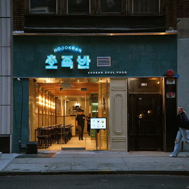 Front view of a Korean restaurant called 'Hojokban' with neon sign in Korean characters and English text 'Korean Soul Food'. The entrance is open, showing a brightly lit interior with tables and a person standing inside. A woman is walking past on the sidewalk.