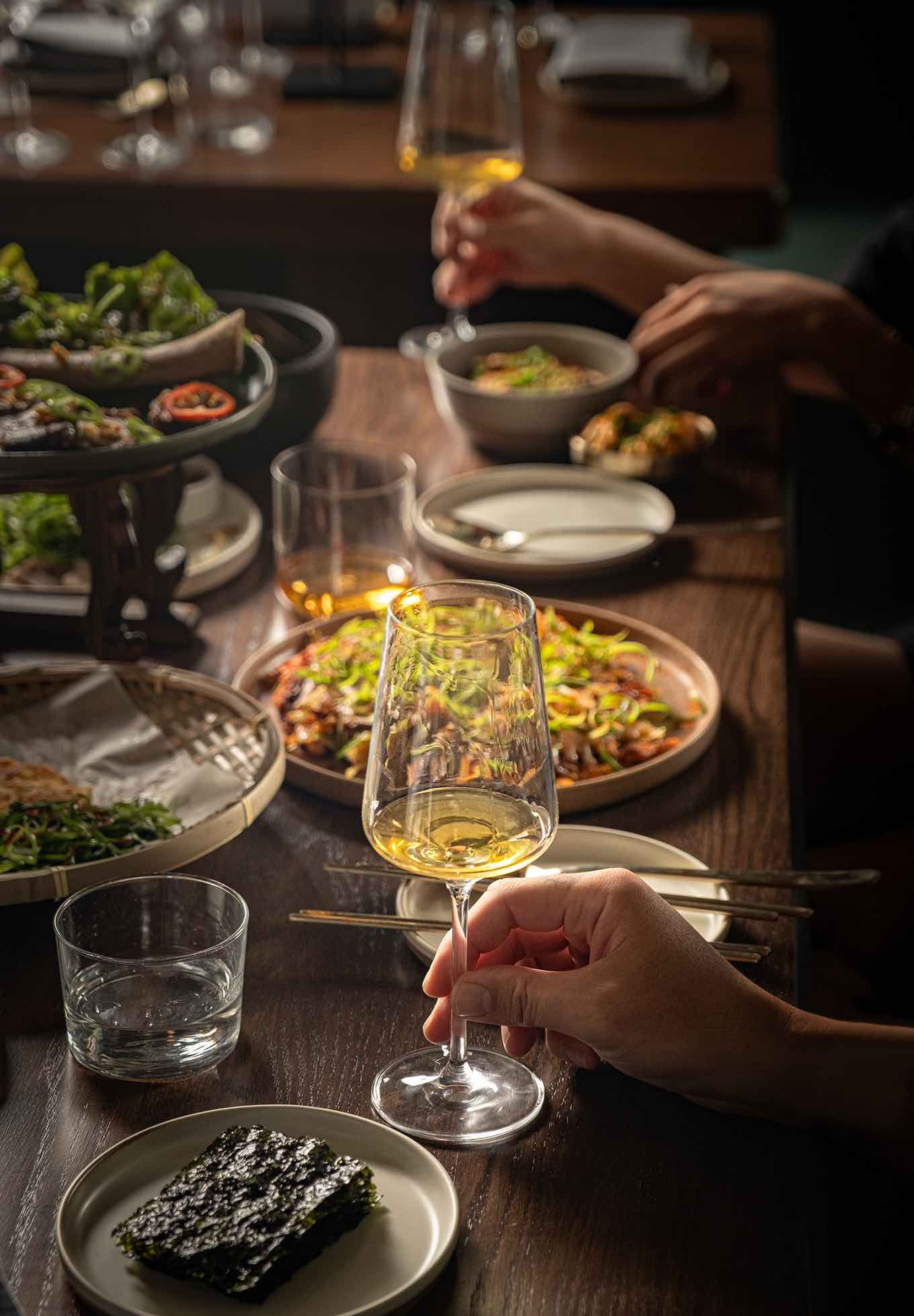 Hand holding a glass of white wine over a shared Korean dining table with assorted dishes at Hojokban in the Downtown LA Arts District