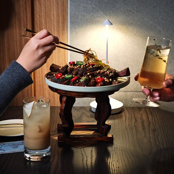 Two people dining with grilled meat and chili peppers, one holding chopsticks and the other holding a glass of beer, on a wooden table with a lit lamp in the background.