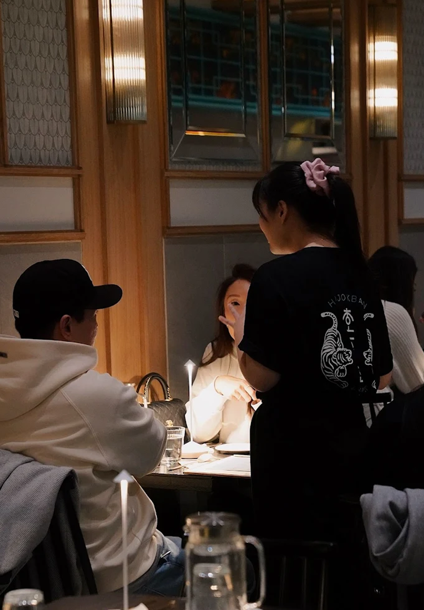 People dining in a restaurant, with one person talking to a woman seated at a table, illuminated by candlelight.