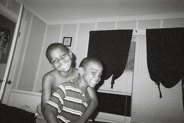 Two young boys smiling and hugging each other in a room with closed curtains.