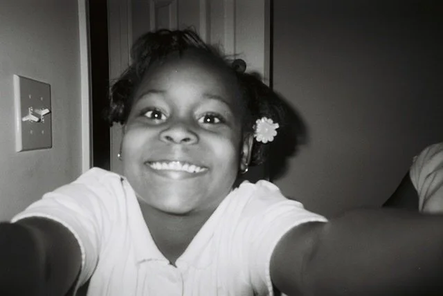 A smiling young girl with a flower in her hair taking a selfie in black and white.