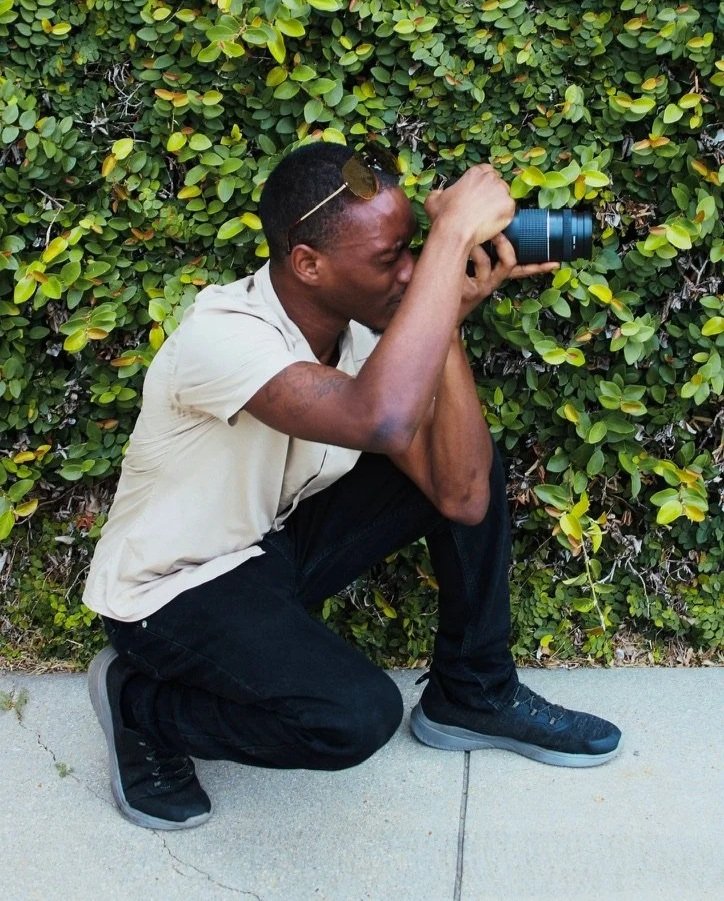 A man crouching on a sidewalk taking a photograph with a camera, with green leafy bushes in the background.
