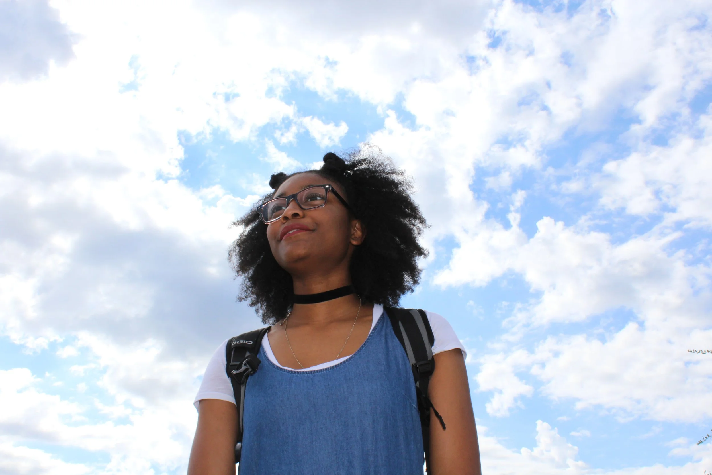 A young woman standing outdoors with a sky filled with clouds in the background.