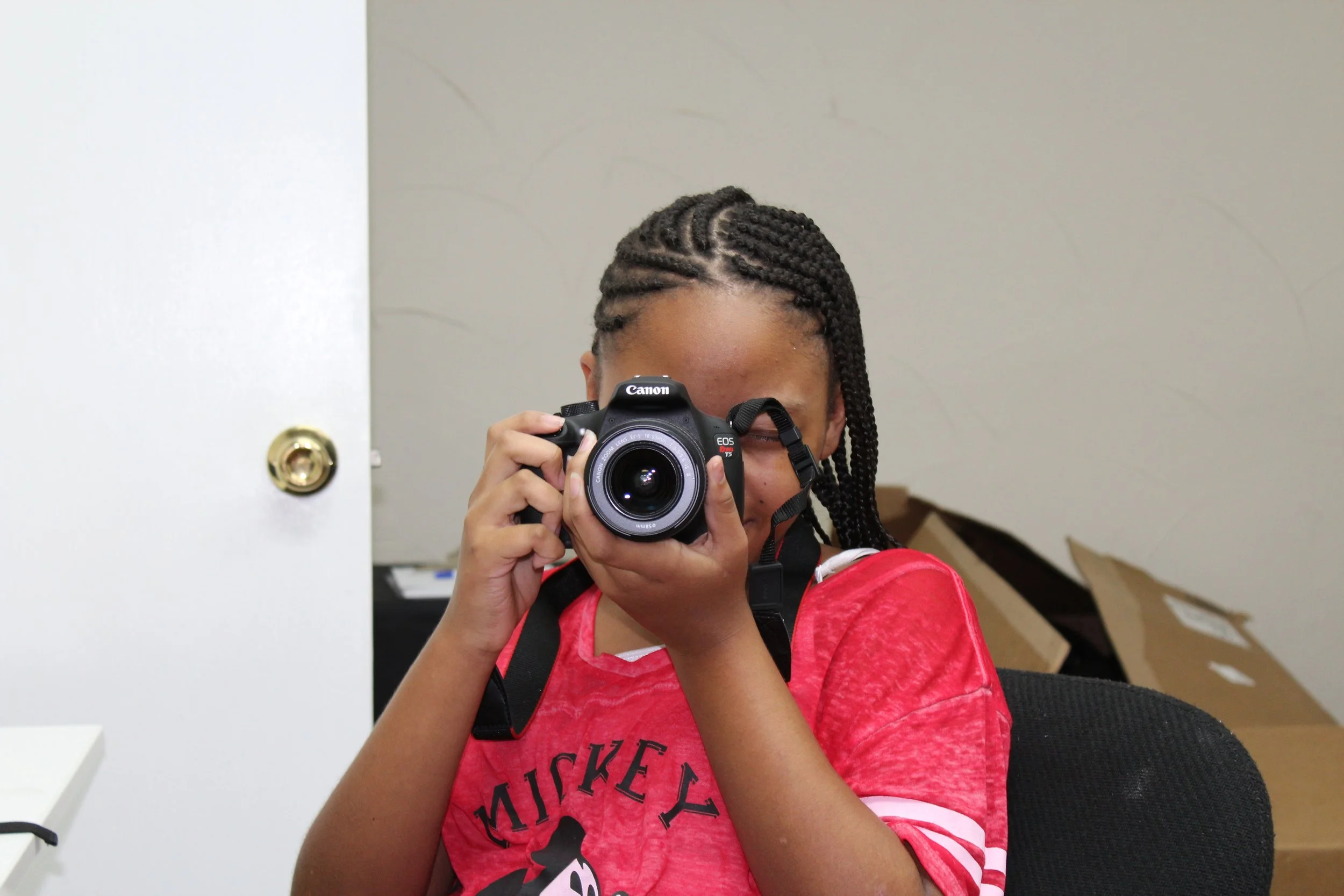 A person with braided hair taking a photo with a Canon camera, wearing a red Mickey Mouse shirt, sitting in a room with cardboard boxes.