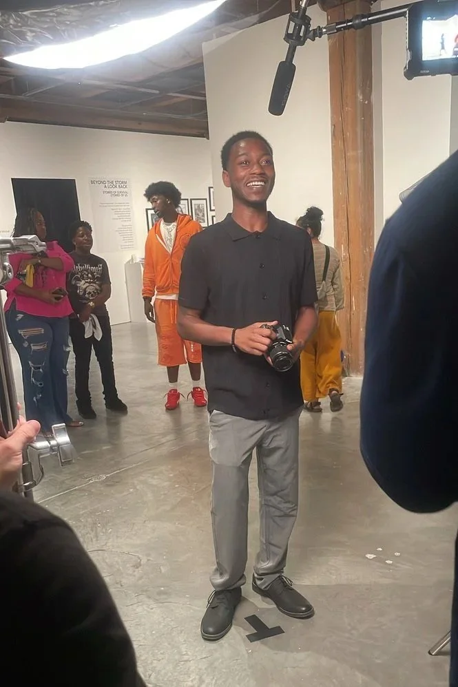 A young man with a camera standing in an art gallery or museum, smiling at the camera, with other visitors and display walls in the background.