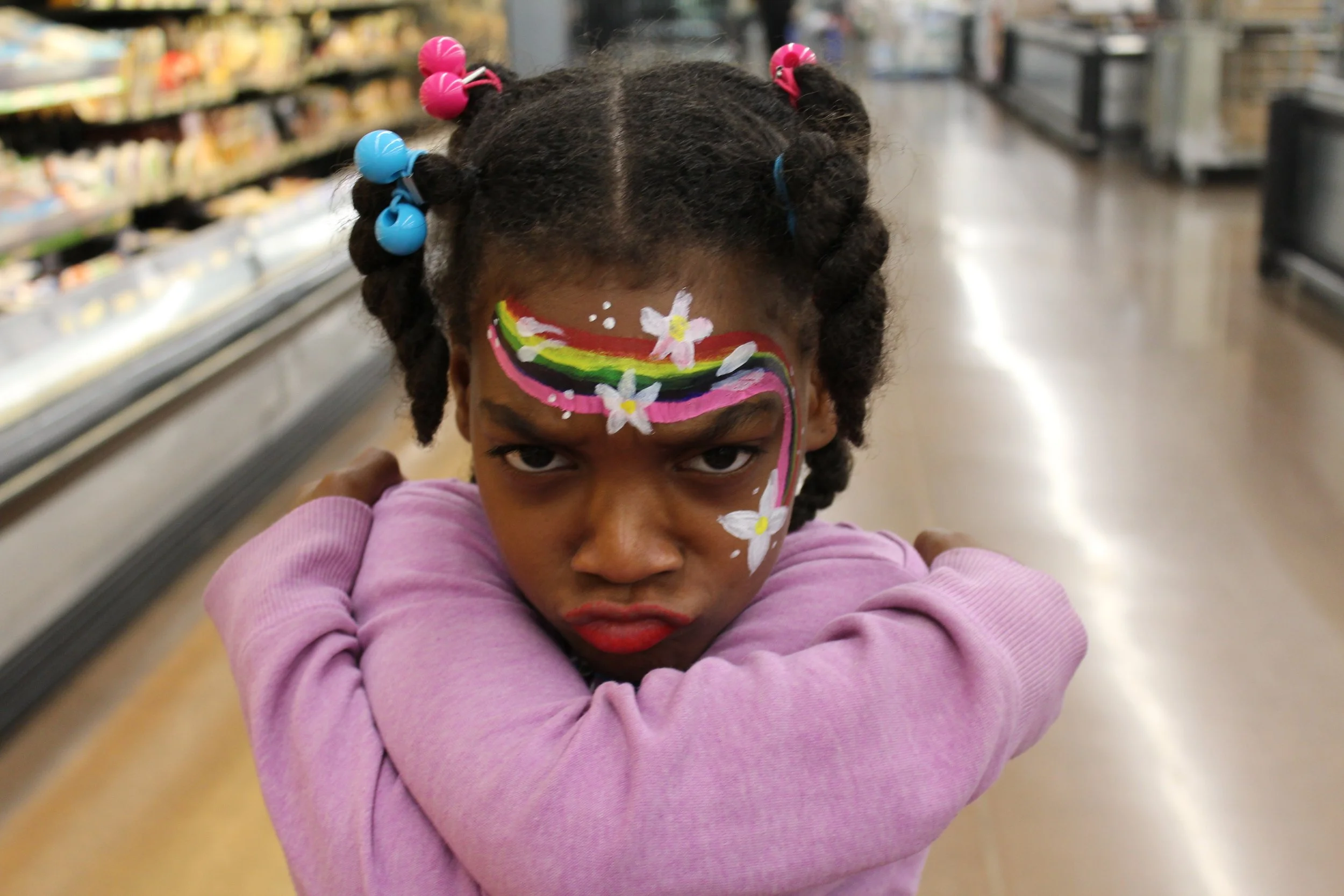 A young girl with face paint of a colorful rainbow with flowers on her forehead in a store aisle, wearing a pink long sleeve shirt, making a fierce expression.