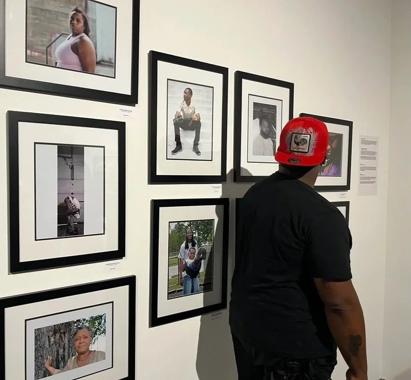 A man wearing a red cap and black shirt is looking at black and white framed photographs on a gallery wall, with some color photos nearby.