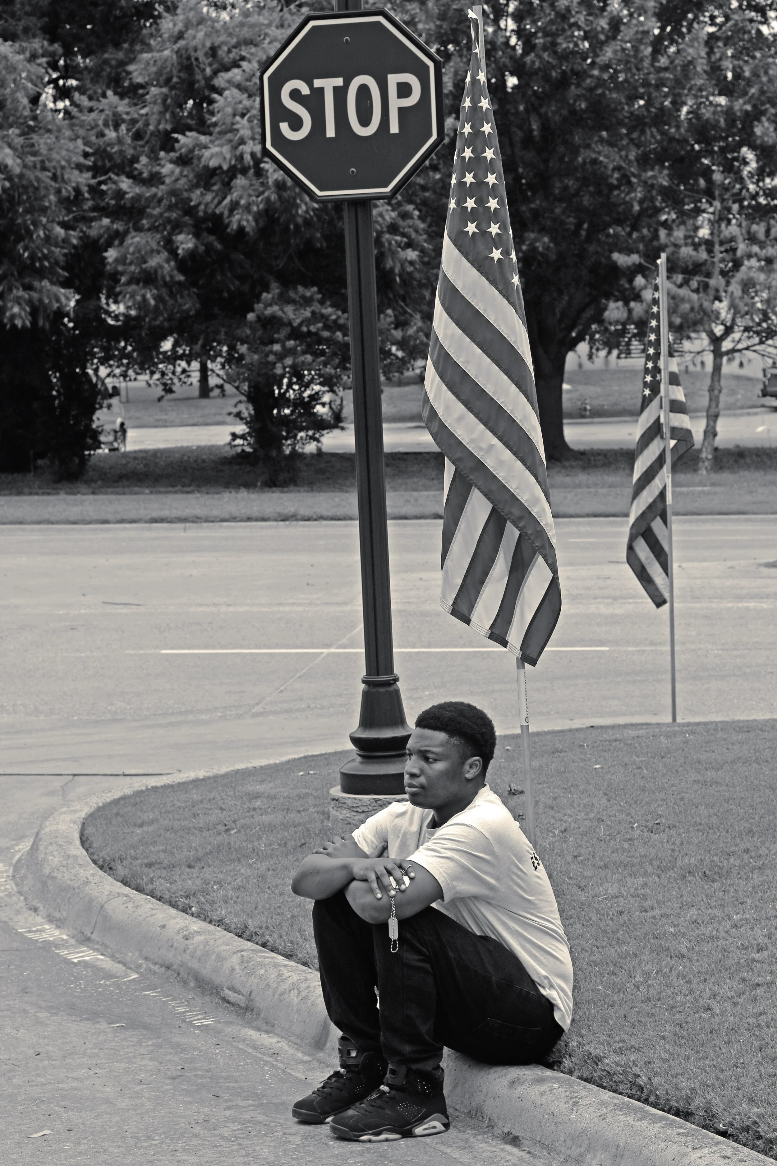 A young man sits on the curb near a stop sign and American flags in a park or yard, with trees and a street in the background.