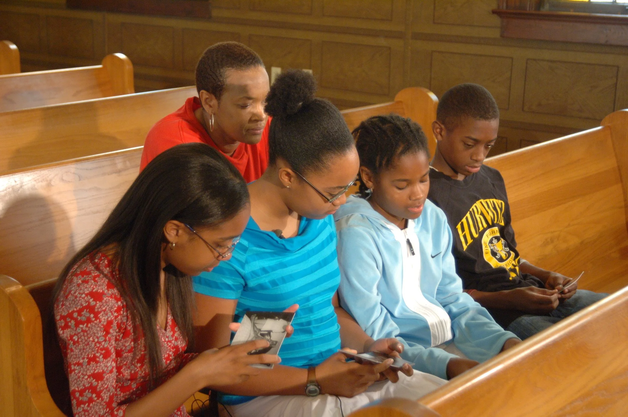 Four Youth Stop Inc. students sitting in church pews looking at printed photos, with Danette Vincent, Founder and Director, sitting behind them answering questions.