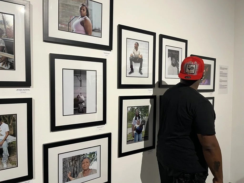 A man wearing a red cap and black shirt is looking at black and white framed photographs on a gallery wall, with some color photos nearby.