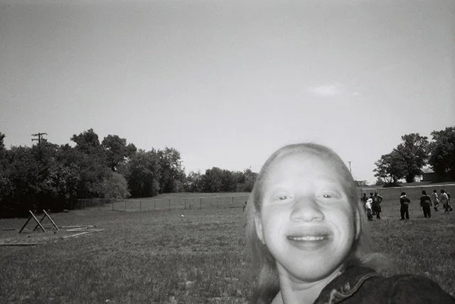 A young girl taking a selfie in a grassy field with trees and a group of people in the background.